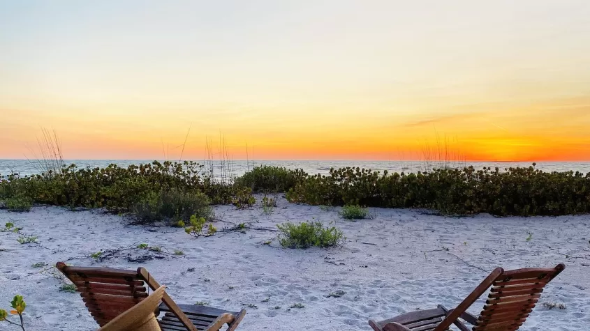 two empty wooden beach chairs facing the sand on the beach and sunset in the background