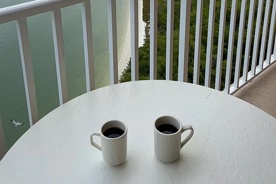 two coffee mugs sitting on an outdoor table on a balcony overlooking water and sunset
